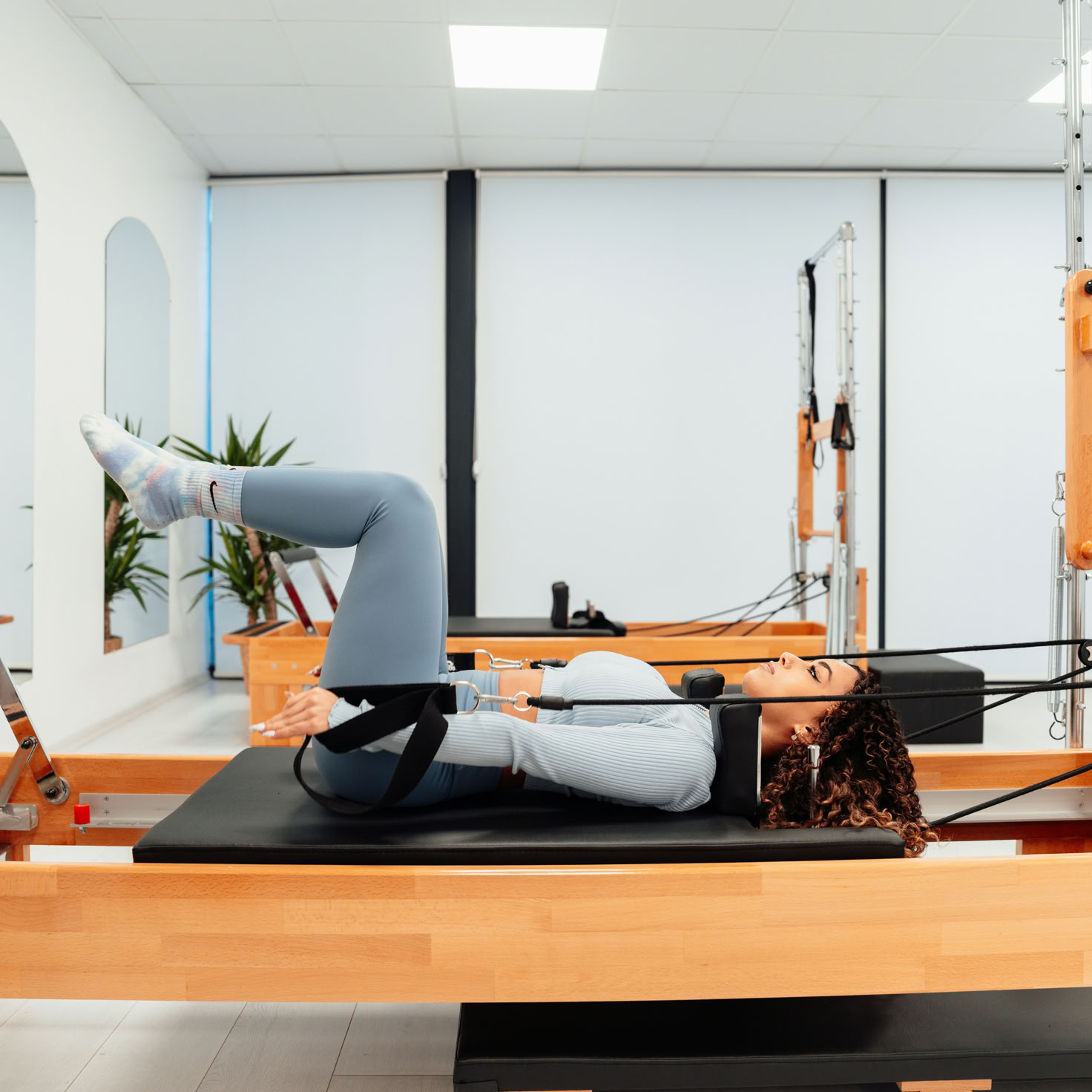 Femme aux cheveux longs roux faisant des exercices sur un appareil de Pilates dans une salle de sport lumineuse.