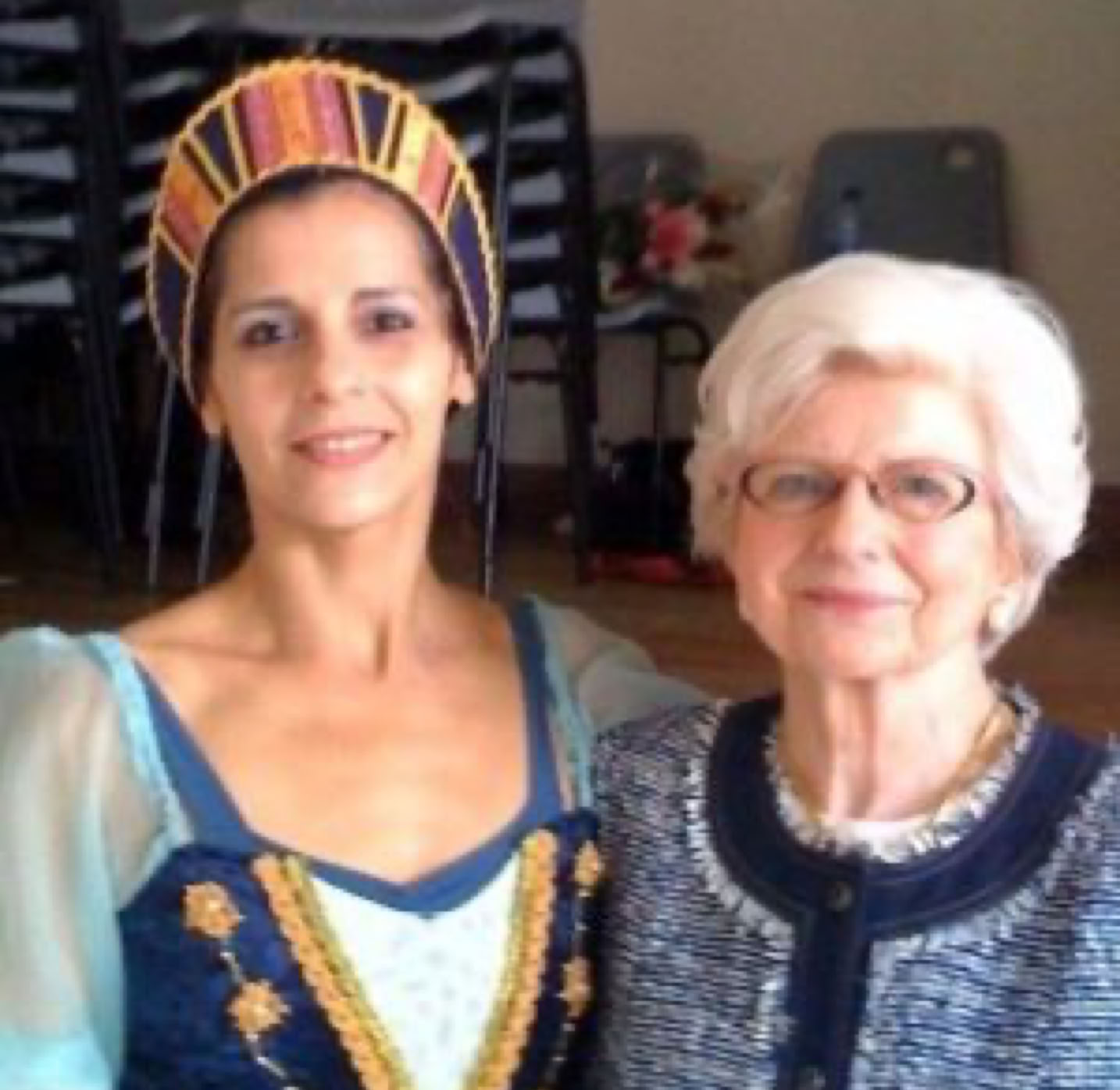 Deux femmes posent ensemble, l'une portant un costume traditionnel coloré et l'autre portant des lunettes et un cardigan bleu.