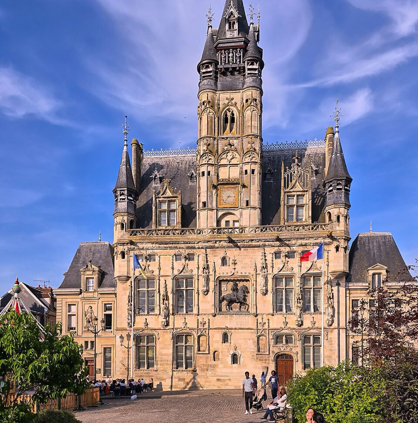 Mairie de Compiègne, bâtiment historique avec une grande tour centrale et des toits en ardoise, entouré de bâtiments plus petits sous un ciel bleu avec des nuages.