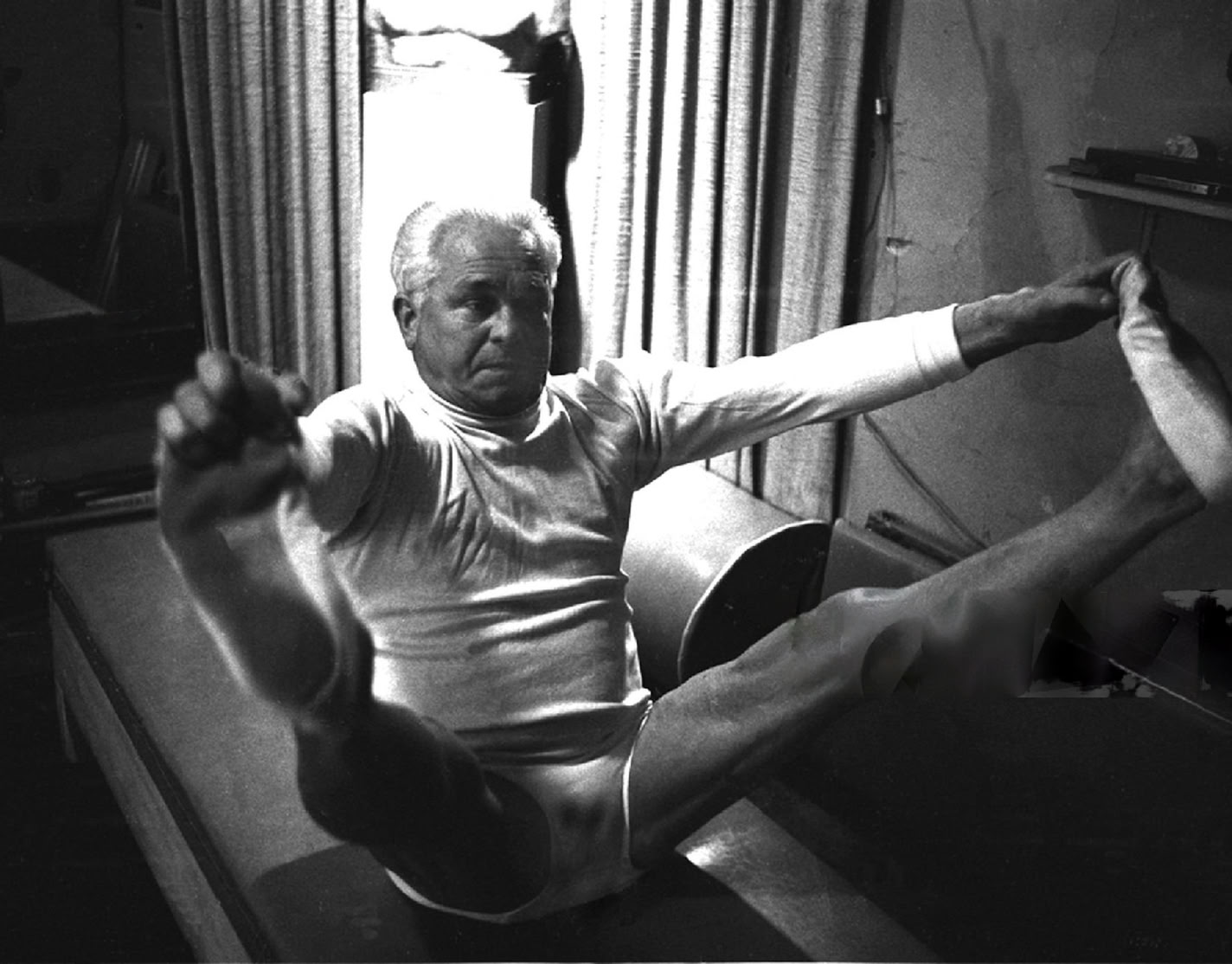 Joseph Pilates allongée sur un appareil de Pilates, les bras tendus vers le haut, dans une pièce avec des fenêtres en arrière-plan.