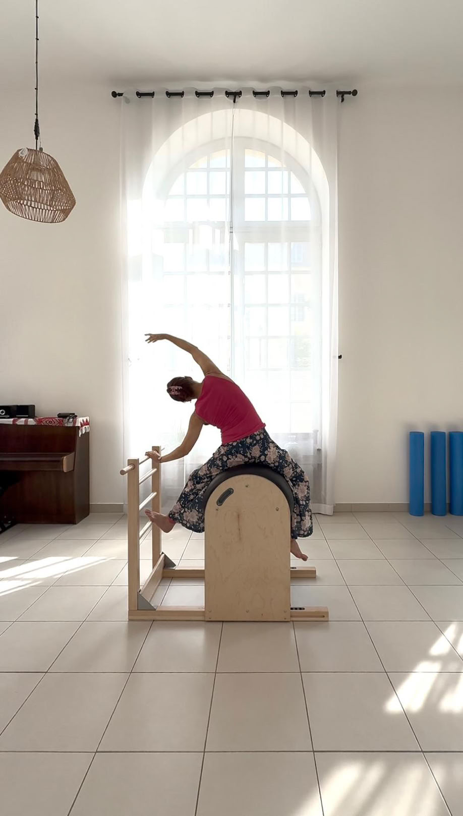 Personne faisant des exercices sur un appareil de Pilates (Barrel) dans une pièce lumineuse avec un grand rideau blanc.