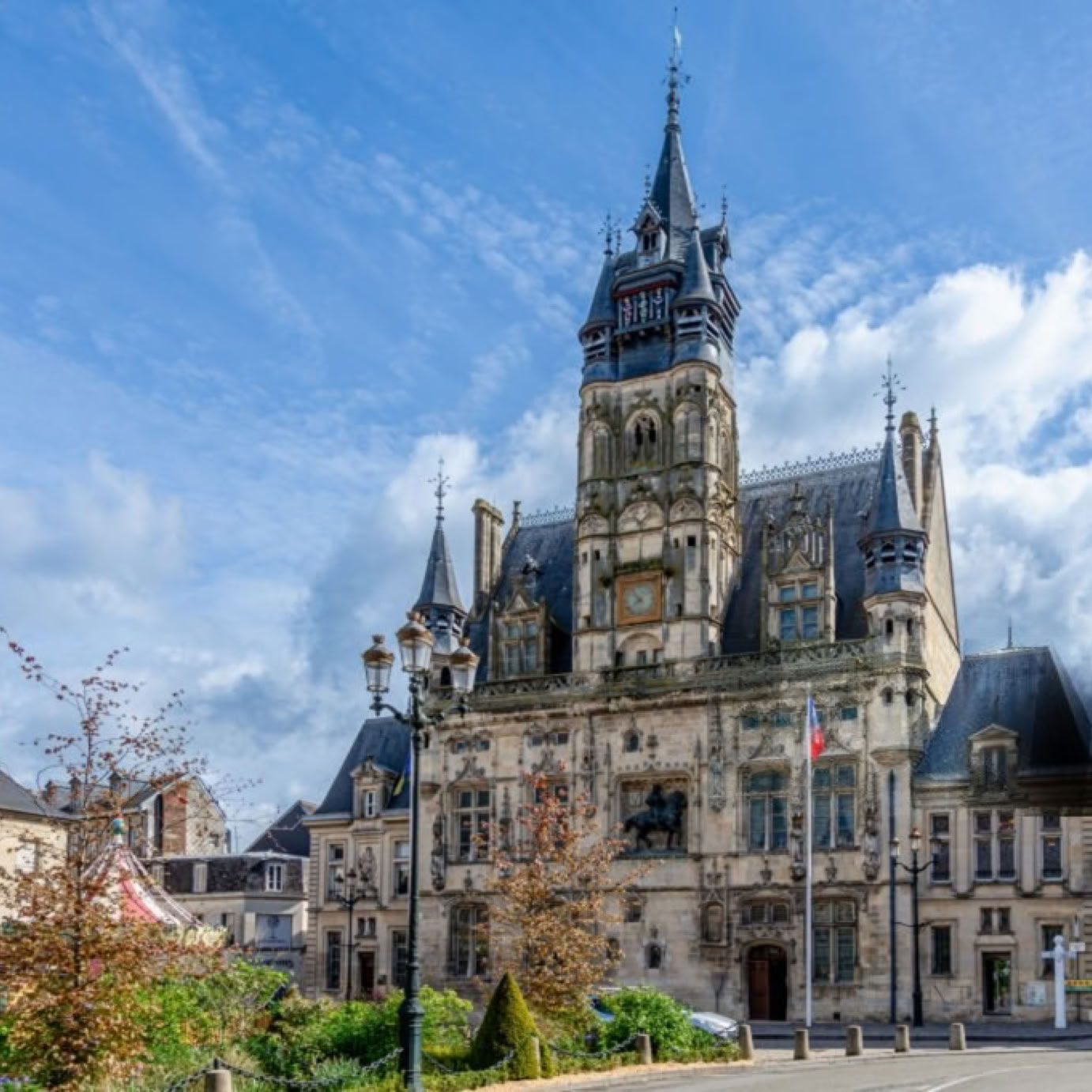 Mairie de Compiègne, bâtiment historique avec une grande tour centrale et des toits en ardoise, entouré de bâtiments plus petits sous un ciel bleu avec des nuages.