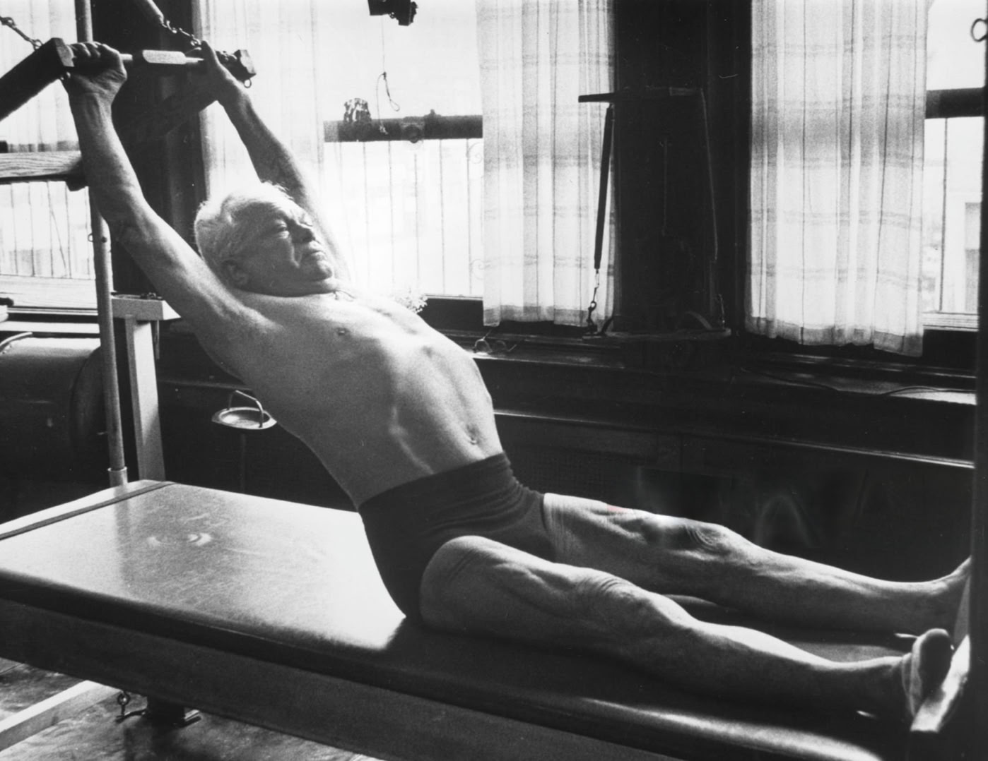 Joseph Pilates allongée sur un appareil de Pilates, les bras tendus vers le haut, dans une pièce avec des fenêtres en arrière-plan.